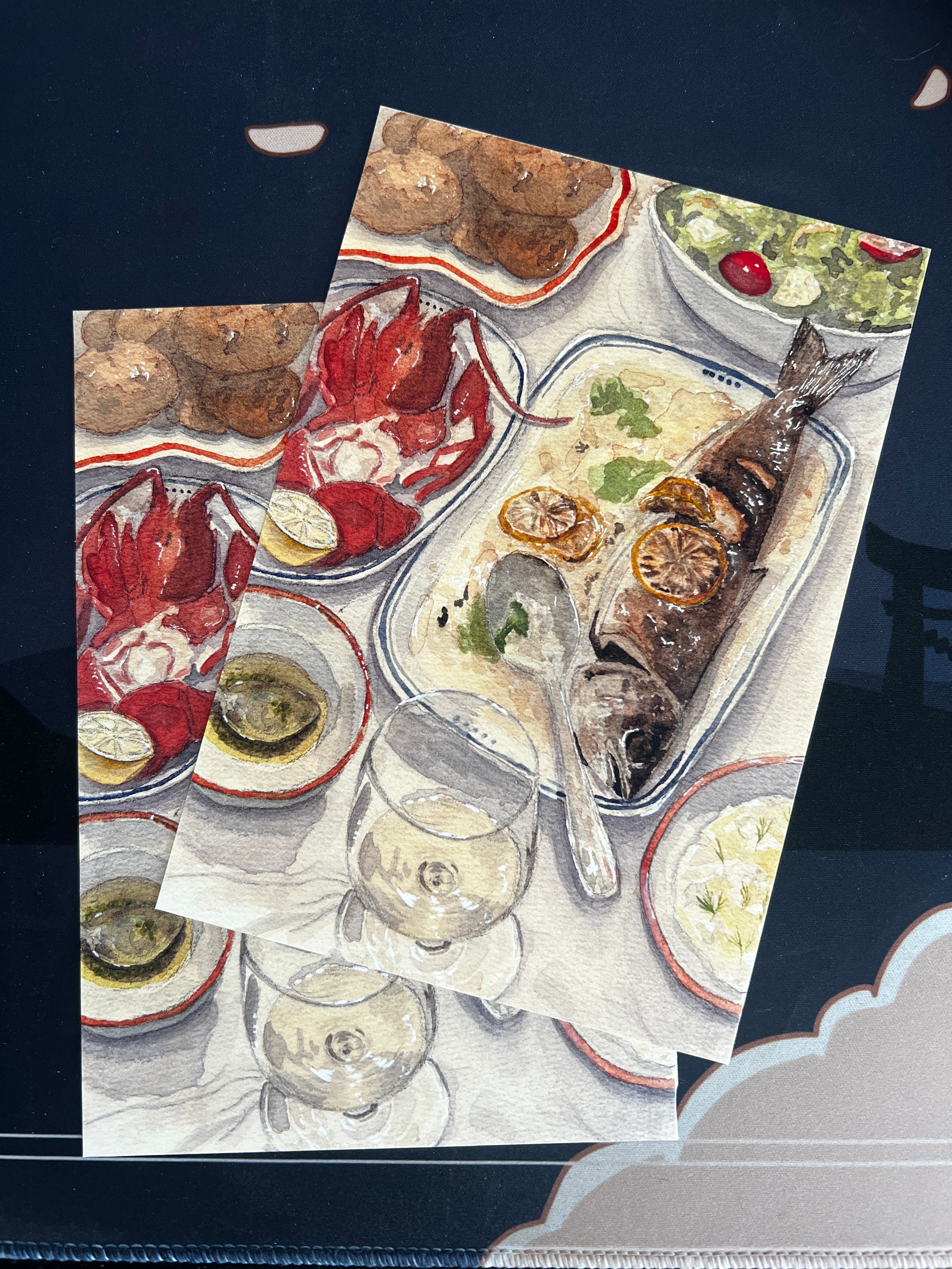 An art print with no borders featuring a painted seafood table scene with a whole grilled fish and lobster, shown overlapping another print against a dark navy background.