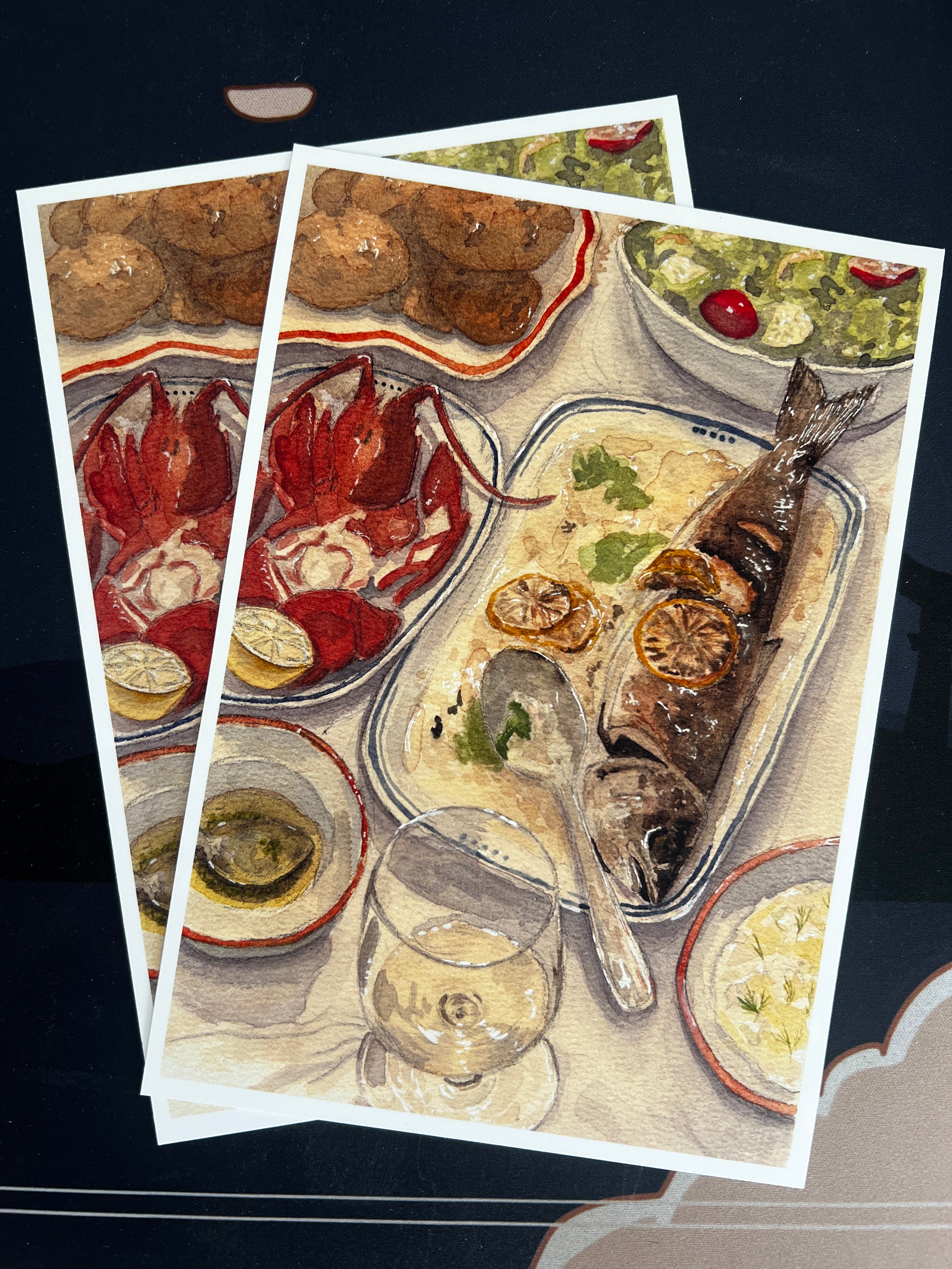 An art print with 1/8" border showing a seafood dinner table scene with a whole fish, lobster, salad, and drinks, photographed against a dark background with overlapping copies visible.